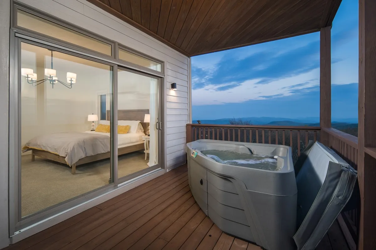 This exterior image captures a bubbling hot tub on a private deck, with views into a cozy bedroom interior and breathtaking mountain scenery beyond. The golden hour lighting suggests evening relaxation, emphasizing both the inviting hot tub and the connection to the bedroom sanctuary.