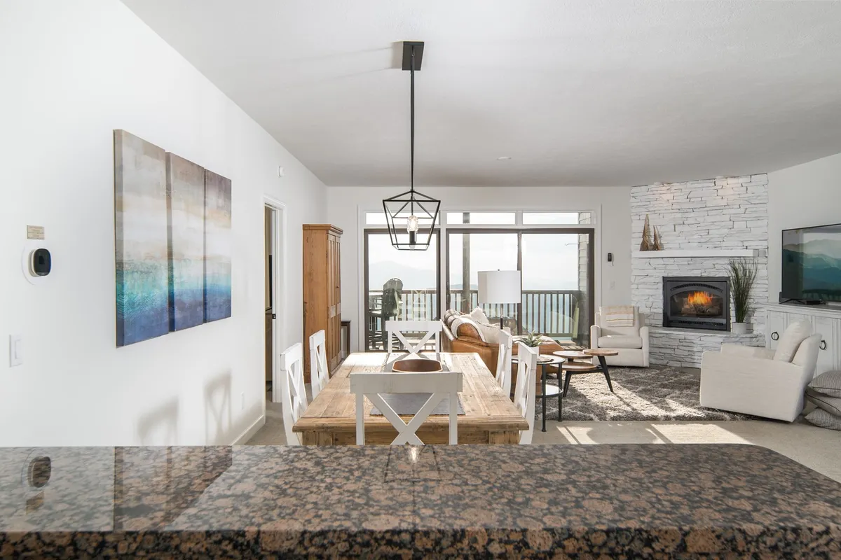 An inviting open-concept living and dining area with a dining table, a fireplace, large windows framing stunning mountain views, and artwork on the wall. The scene evokes a sense of warmth and community, with comfortable seating and abundant natural light.