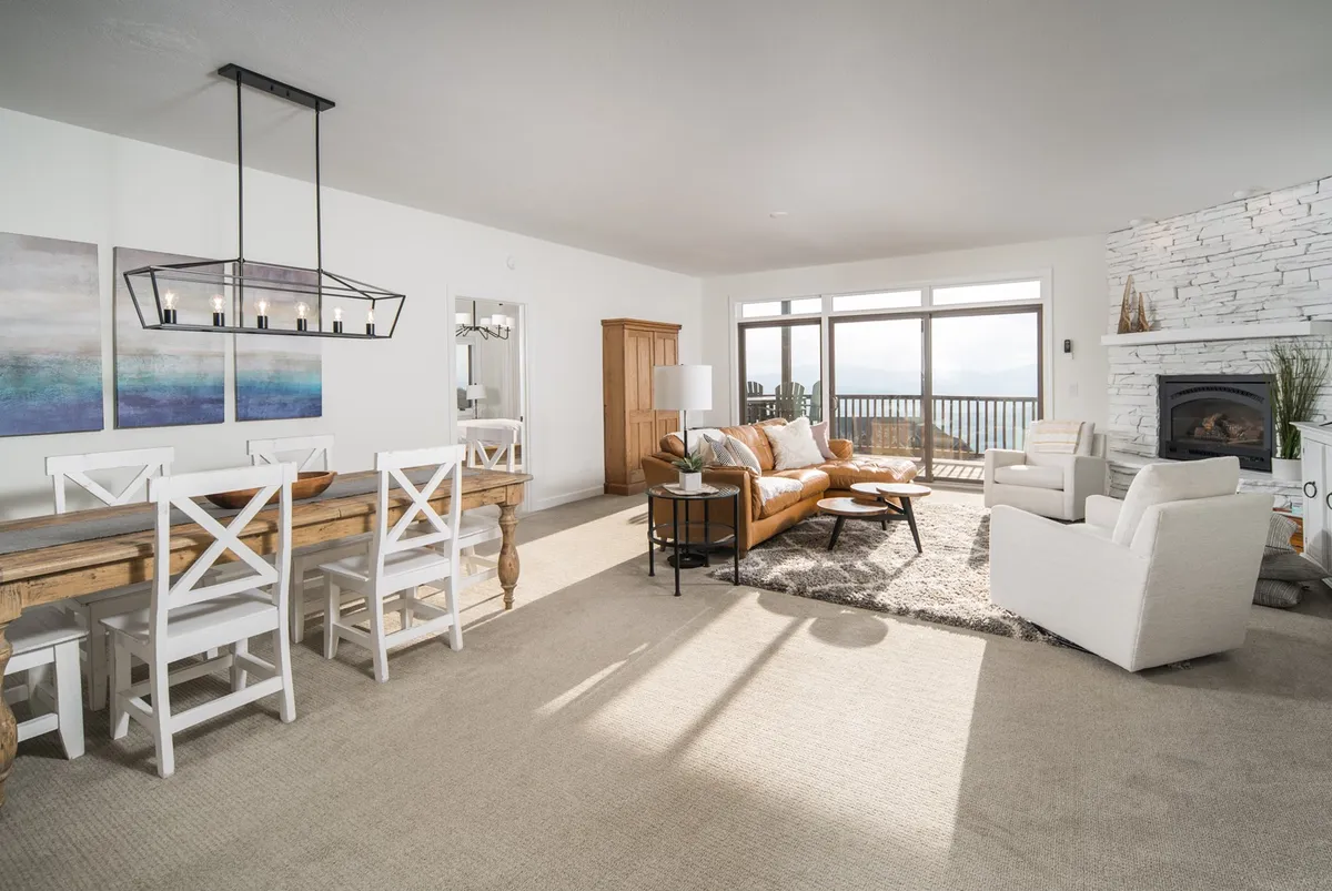 A bright, cozy living space with a stone fireplace, large windows looking out to the mountains, a dining table, and a comfortable seating area. The room is filled with warm tones, inviting both relaxation and social connection by the fire.