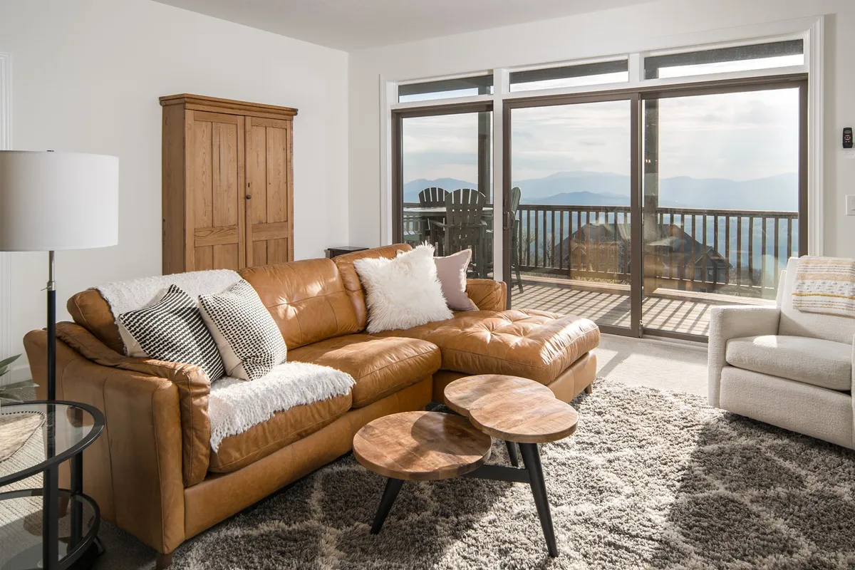 An inviting living space featuring a comfortable leather couch, wooden coffee tables, and large windows framing a scenic mountain view. The room is filled with natural light, and the setup encourages relaxation after a day outdoors.