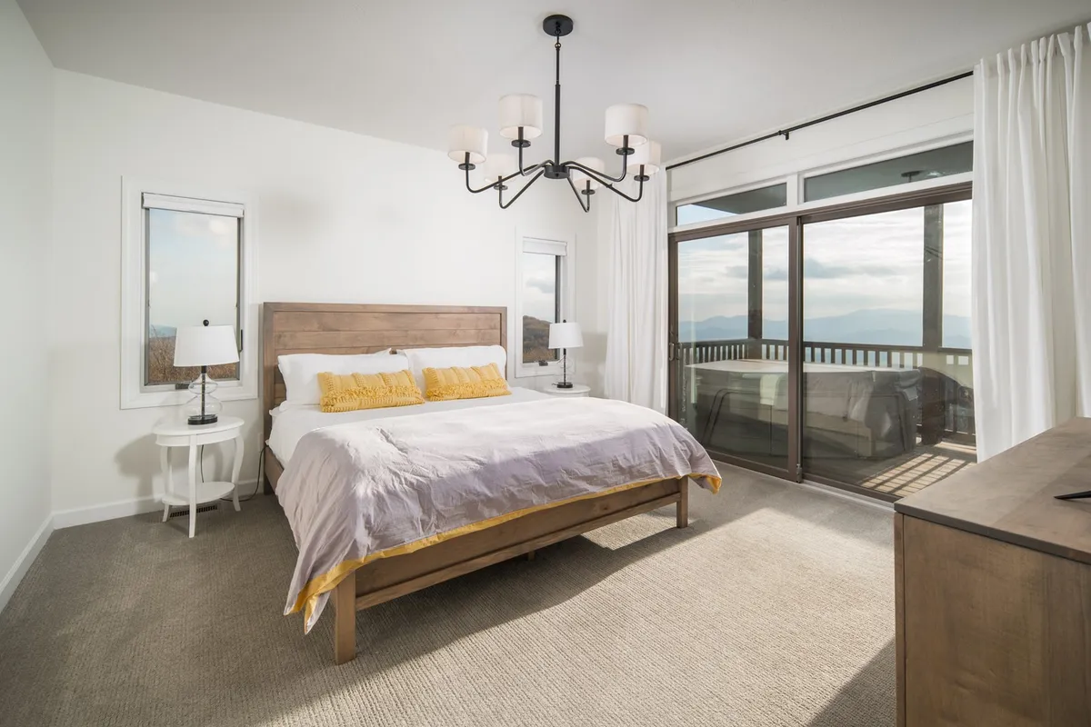 A bright and spacious bedroom featuring a king-sized bed adorned with yellow accent pillows. A stylish chandelier hangs above, while large sliding glass doors let in abundant natural light and offer a view outdoors. Side tables with lamps flank the bed, creating a cozy, inviting atmosphere ideal for relaxation.