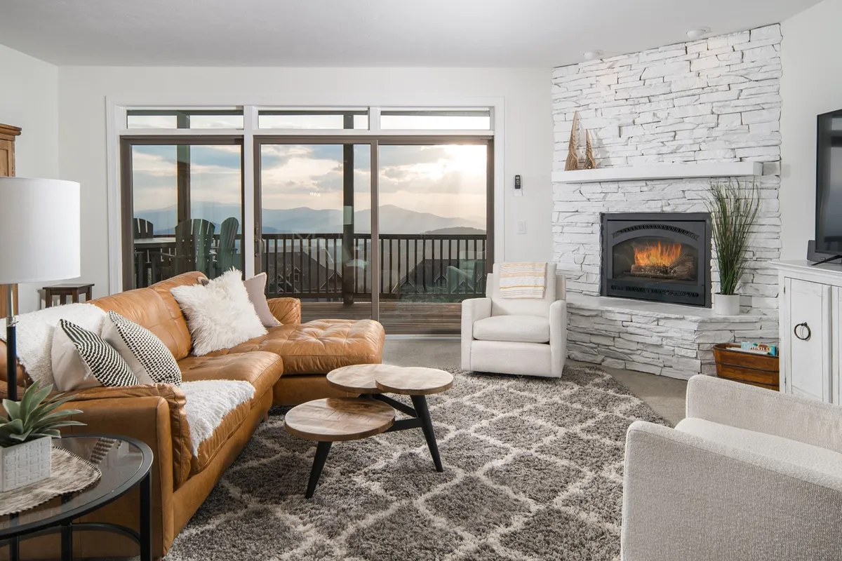 A warm, inviting living room with a plush leather sofa facing a stunning stone fireplace. Floor-to-ceiling windows frame breathtaking mountain vistas, bathing the room in natural light and offering the perfect spot to watch the sunset. The scene is set for a cozy evening with loved ones, enjoying the peaceful ambiance and comforts of a luxury mountain retreat.
