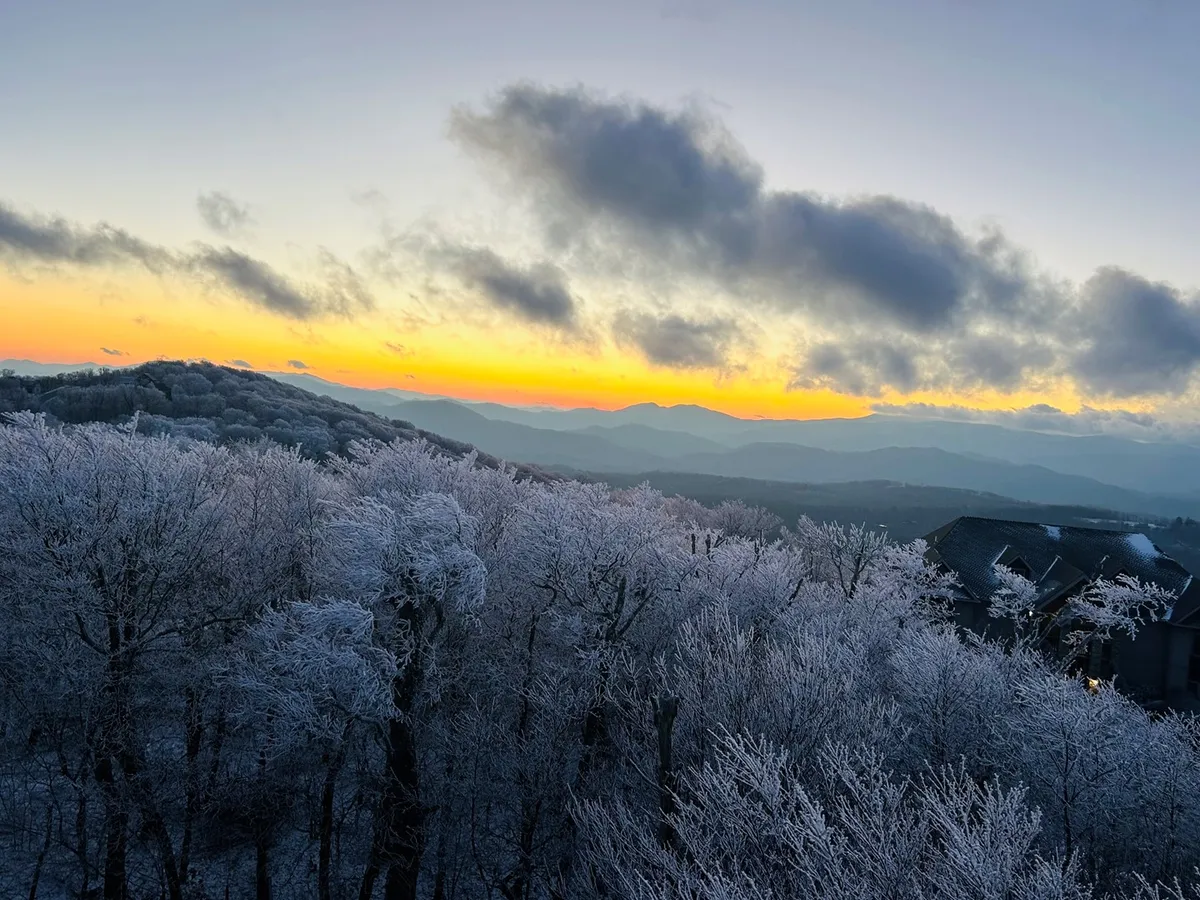 A stunning outdoor view featuring frost-covered trees, a glowing sunset sky, and rugged mountain ranges in the distance. The winter landscape underscores the area's seasonal appeal, hinting at skiing and snowboarding opportunities.