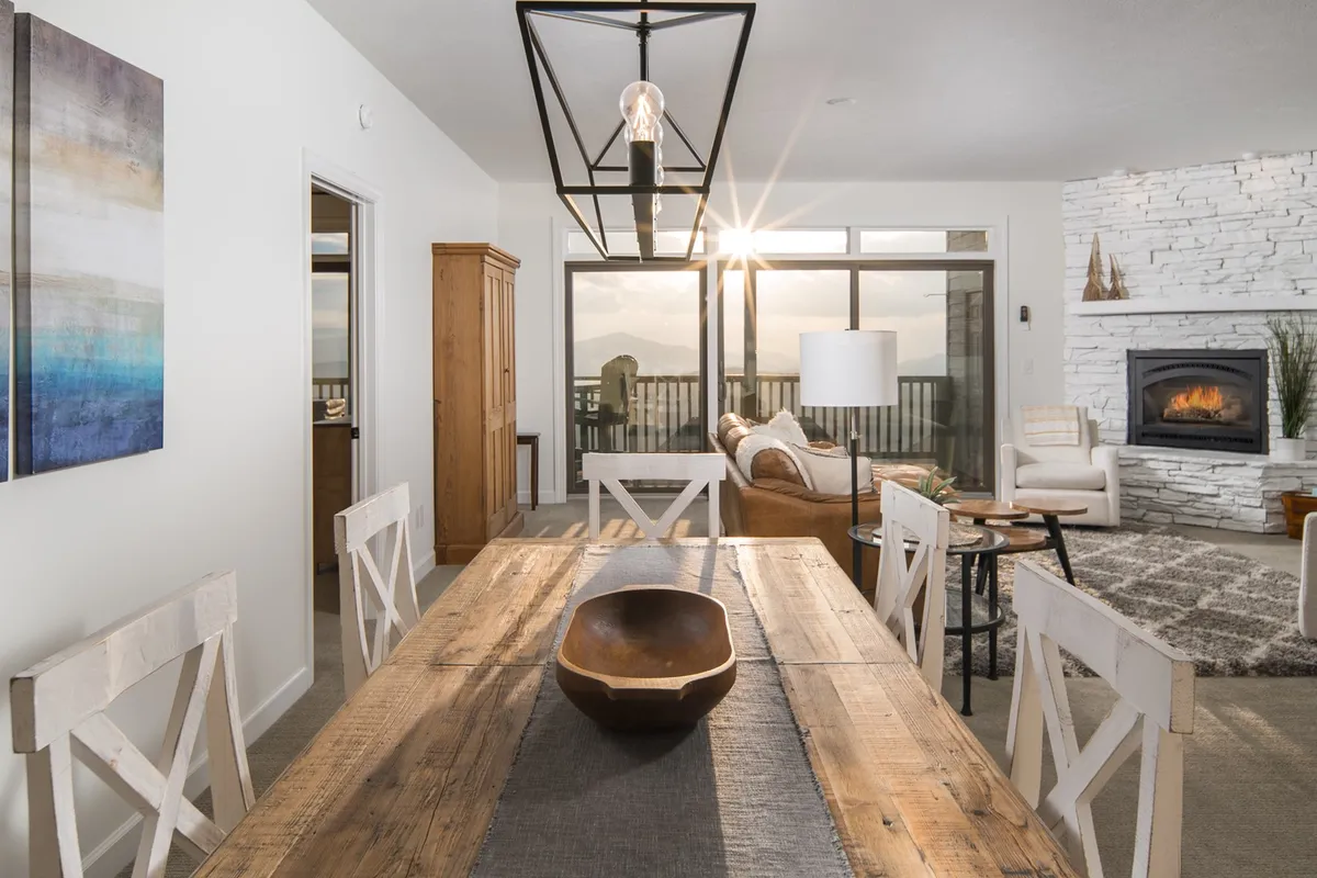 A spacious, open-concept living area featuring a cozy electric fireplace as the focal point. Plush seating surrounds the fireplace, while a large dining table sits nearby. Expansive windows invite natural light and offer sweeping views of the mountains, creating a warm, welcoming atmosphere ideal for gatherings and relaxation.