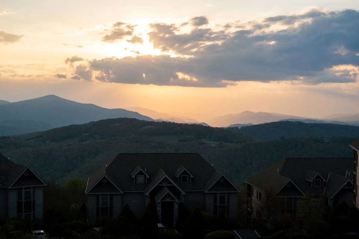 A panoramic view of the Blue Ridge mountains at sunset, with soft golden light illuminating the rolling peaks and valleys. Distant houses nestle into the landscape, emphasizing the tranquility and seclusion of the setting.