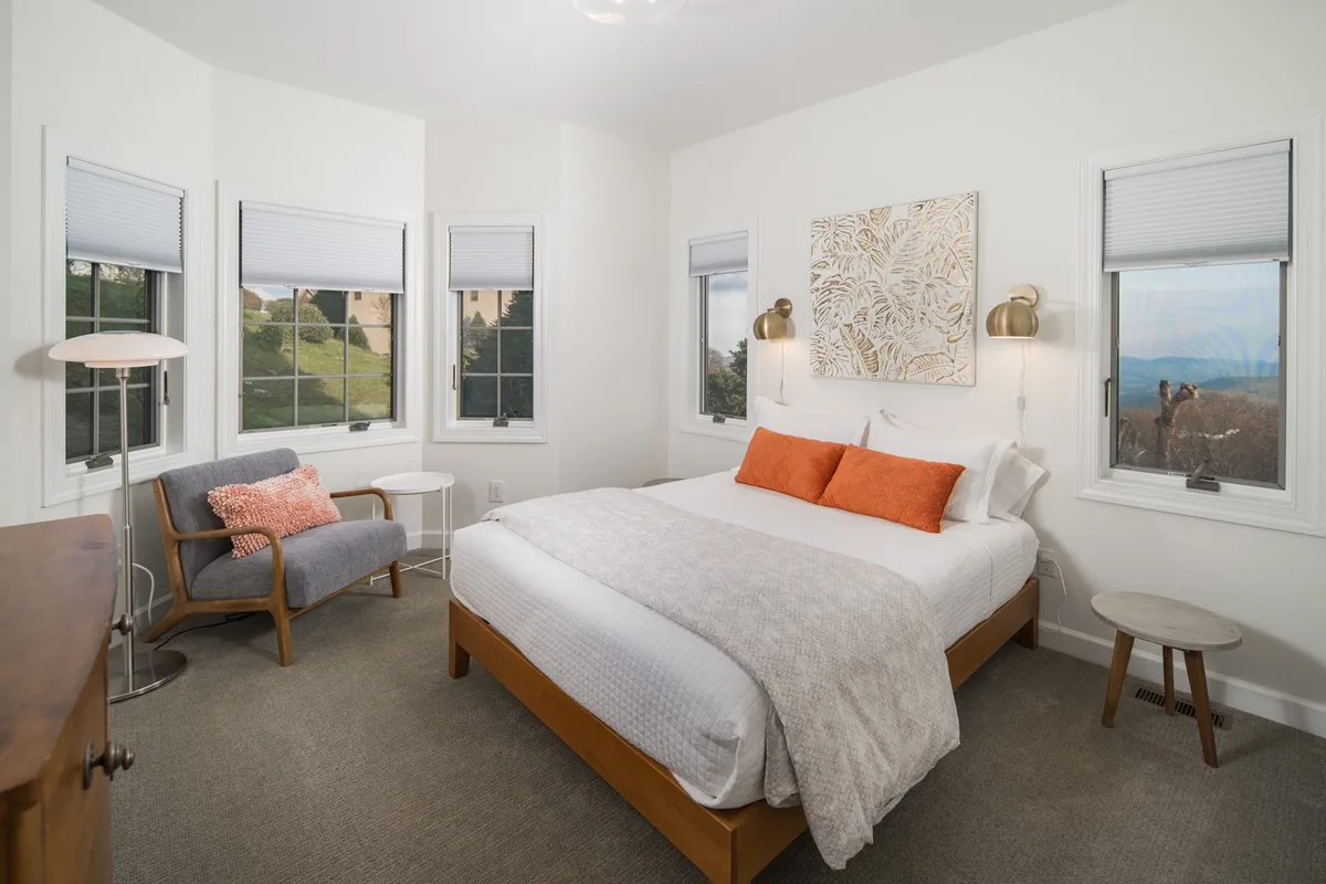 A king-size bed with orange accent pillows forms the focal point of this tranquil bedroom. Artwork decorates the wall, and a cozy armchair sits beside a window, creating a perfect nook for reading or quiet reflection. Soft light streams into the room, enhancing its peaceful vibe.